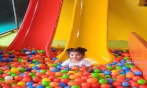 A child enjoying a colorful ball pit with red and yellow slides in an indoor play area, surrounded by vibrant plastic balls.